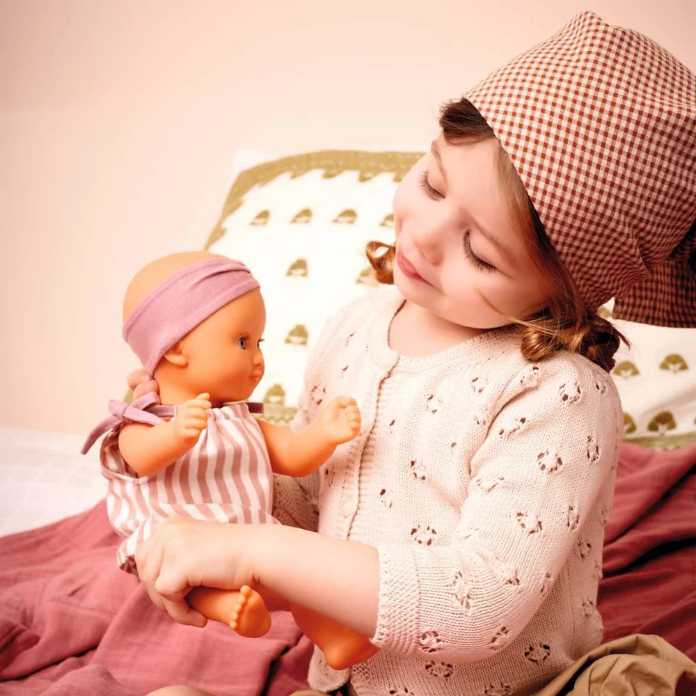 POMEA Badepuppe Mädchen Prune in rosa-weißem Kleid mit Mütze ; POMEA Badepuppe Mädchen Prune mit rosa Badeanzug und Stirnband ; POMEA Badepuppe Baby Prune 32cm mit rosa Outfit und Kopftuch ; POMEA Badepuppe Mädchen Prune liegt auf Wickelunterlage auf Tuch ; Kleines Mädchen mit POMEA Badepuppe in Rosa spielend auf Bett ; Mädchen mit POMEA Badepuppe in rosa Puppenkleidung spielend ; POMEA Badepuppe Mädchen Prune mit Zubehör beim Baden spielen