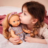 POMEA Puppe Lovely Marie im Originalkarton mit Blumenkleid ; Kleines Mädchen küsst POMEA Puppe Lovely Marie mit blondem Haar ; Pomea Puppe Lovely Marie mit blondem Haar und Blumenkleid von Djeco ; POMEA Puppe Lovely Marie mit blondem Haar, rosa Haarband und Blumenkleid ; POMEA Puppe Lovely Marie mit goldblondem Haar von hinten ; POMEA Puppe Lovely Marie mit Holzkleiderstander und Puppenkleidung ; Kind spielt mit POMEA Puppe Lovely Marie und Stethoskop