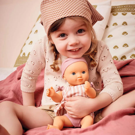 POMEA Badepuppe Mädchen Prune in rosa-weißem Kleid mit Mütze ; POMEA Badepuppe Mädchen Prune mit rosa Badeanzug und Stirnband ; POMEA Badepuppe Baby Prune 32cm mit rosa Outfit und Kopftuch ; POMEA Badepuppe Mädchen Prune liegt auf Wickelunterlage auf Tuch ; Kleines Mädchen mit POMEA Badepuppe in Rosa spielend auf Bett ; Mädchen mit POMEA Badepuppe in rosa Puppenkleidung spielend ; POMEA Badepuppe Mädchen Prune mit Zubehör beim Baden spielen